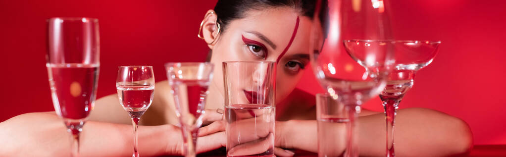 asiatische Frau mit künstlerischem Aussehen und Ohrenmanschette, die in die Kamera schaut, nahe einer Brille mit reinem Wasser auf rotem Grund, Banner - Foto, Bild