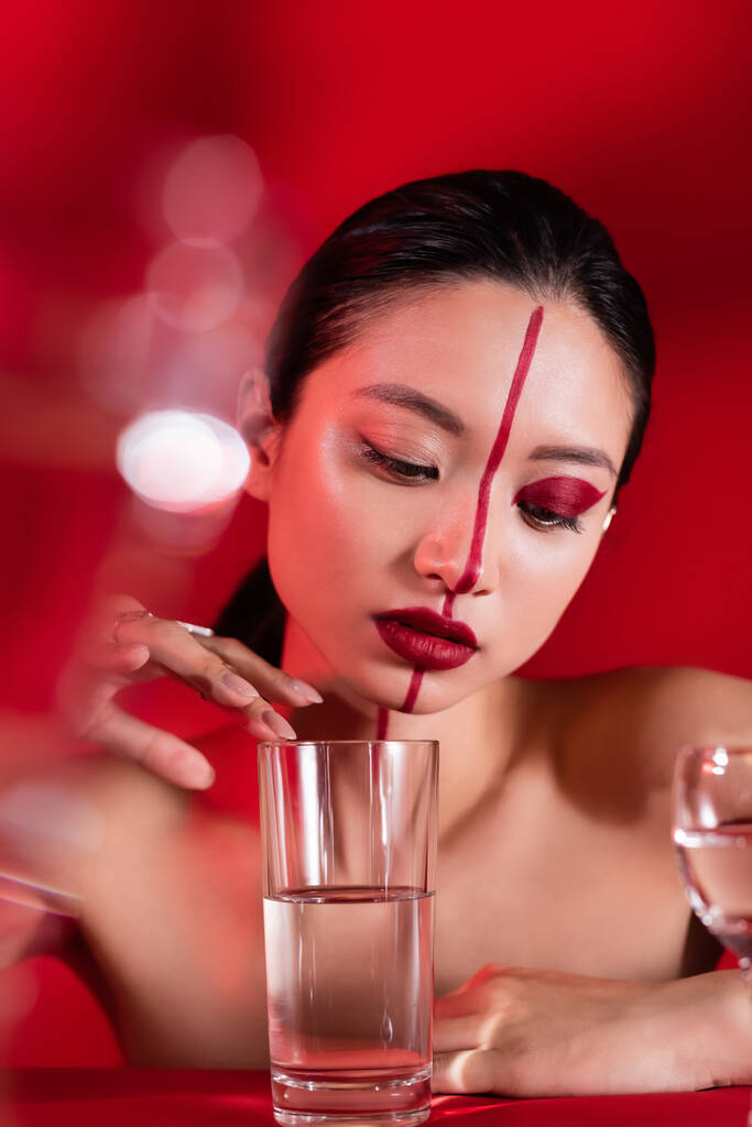 jovem mulher asiática com maquiagem criativa e ombros nus tocando vidro com água pura no fundo vermelho - Foto, Imagem
