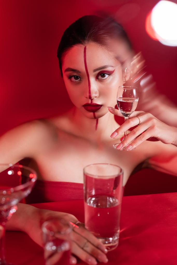 brunette asian woman with red creative visage holding glass of pure water on blurred foreground - Photo, Image
