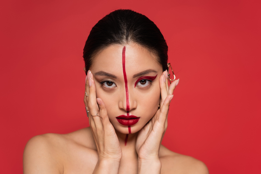 brunette asiatique femme avec les épaules nues et artistique maquillage tenant les mains près du visage isolé sur rouge - Photo, image