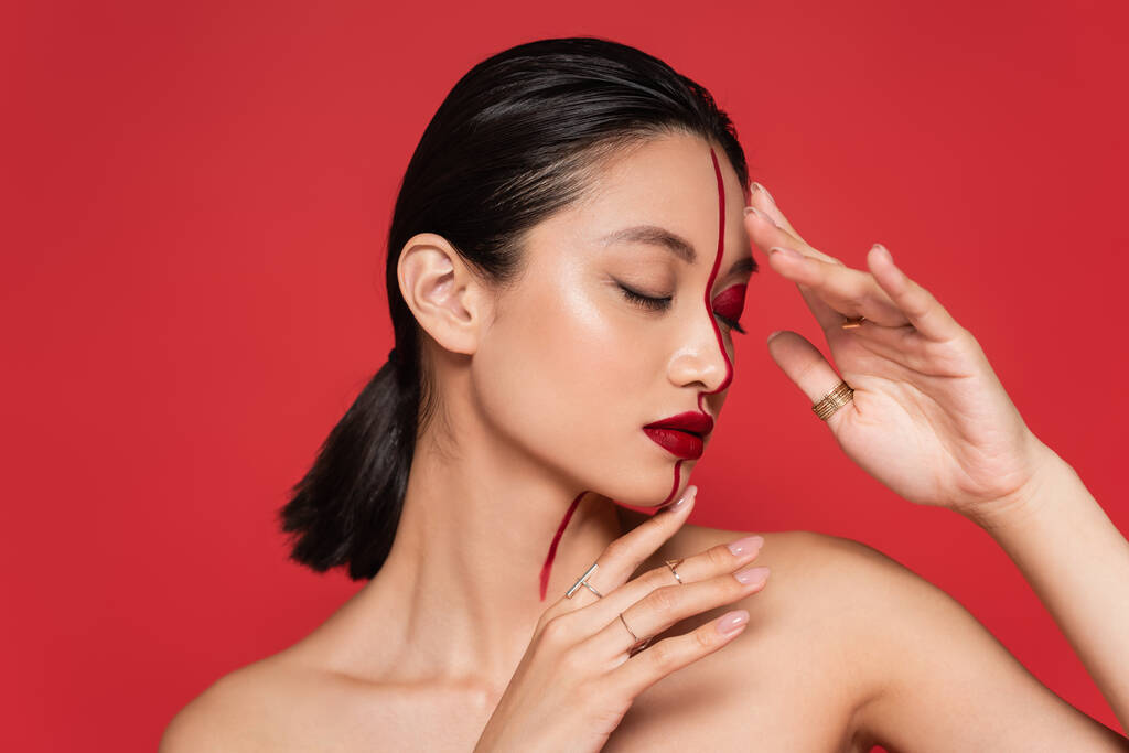 чувственная азиатская женщина с голыми плечами и закрытыми глазами, держась за руки рядом с лицом с творческим макияжем, изолированным на красном - Фото, изображение