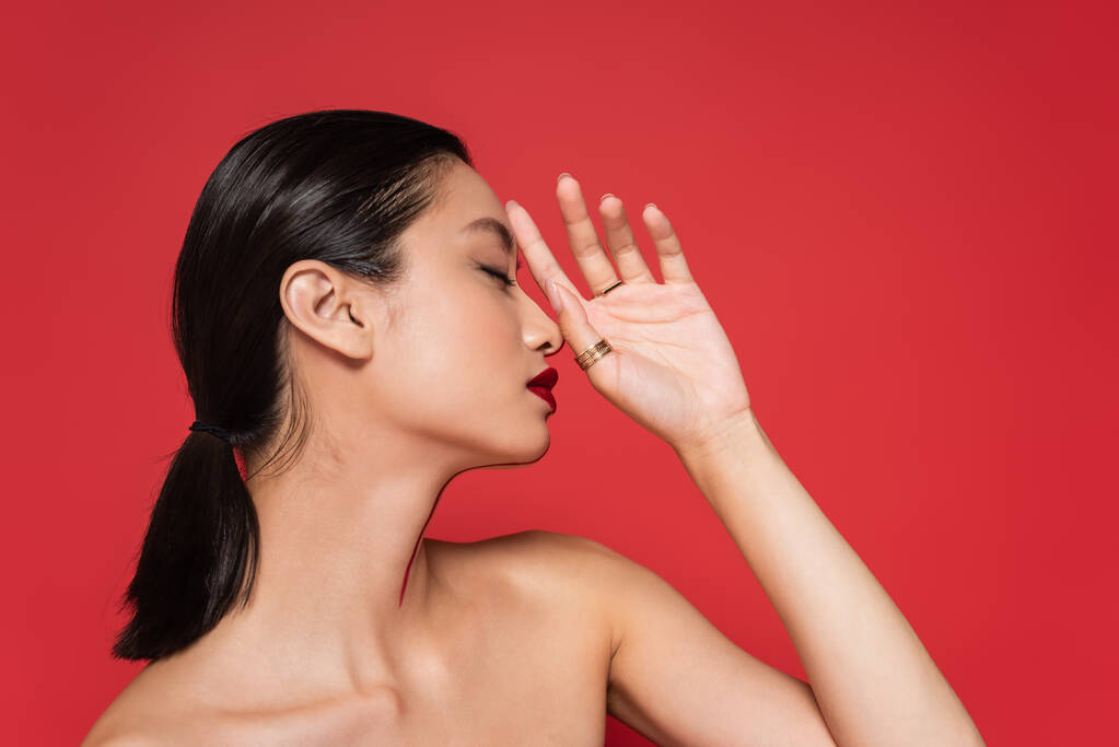 profil brunetka azjatyckie kobieta z nagie ramiona i makijaż pozowanie z ręka blisko twarz odizolowany na czerwony - Zdjęcie, obraz