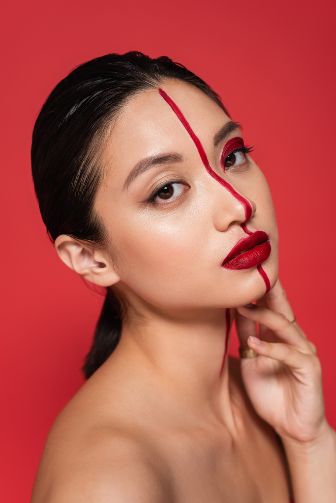 portret van aziatisch model met creatief gelaat en perfecte huid hand in hand in de buurt van gezicht en kijken naar camera geïsoleerd op rood - Foto, afbeelding
