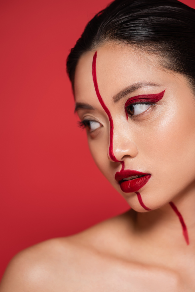 portrait of sensual asian woman with bare shoulder and creative makeup looking away isolated on red - Photo, Image