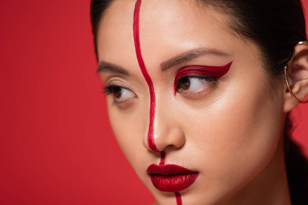γκρο πλαν πορτρέτο της Ασιάτισσας με καλλιτεχνικό μακιγιάζ στο πρόσωπο χωρισμένο με γραμμή που απομονώνεται στο κόκκινο - Φωτογραφία, εικόνα