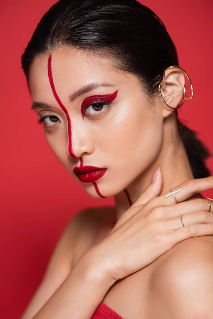 портрет соблазнительной азиатки с манжетой уха и ярким художественным обликом, касающимся обнаженного плеча, изолированного на красном - Фото, изображение