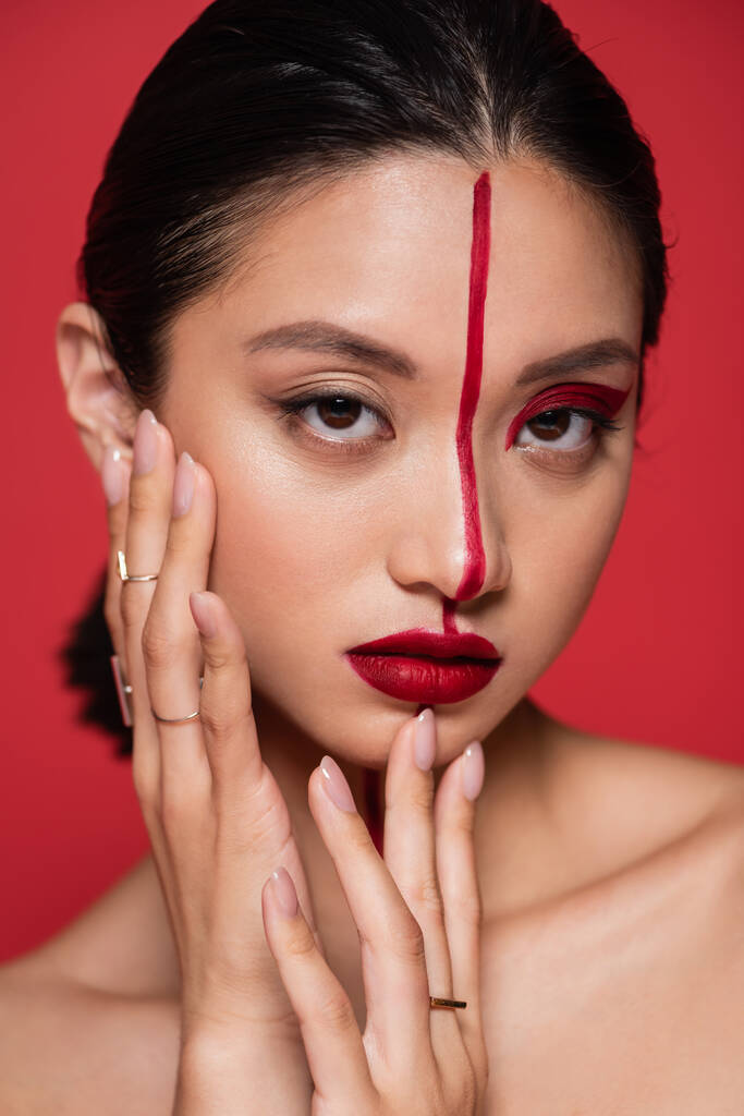 portret van aziatische vrouw aanraken perfect gezicht met creatieve make-up en kijken naar camera geïsoleerd op rood - Foto, afbeelding