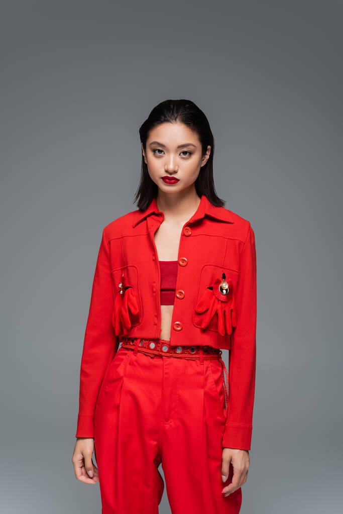молодая азиатка в красной куртке, украшенной брошками и перчатками, смотрящая на камеру, изолированную на сером - Фото, изображение