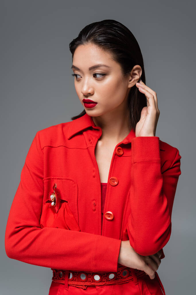 mujer asiática de moda en chaqueta roja tocando el pelo morena y mirando a otro lado aislado en gris - Foto, imagen