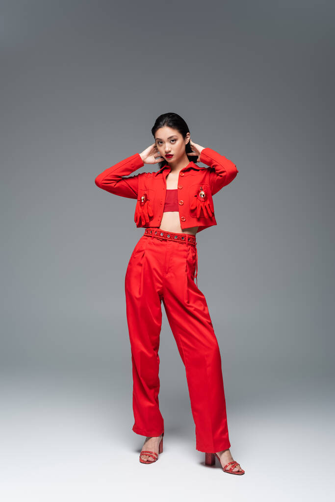 полная длина азиатской женщины в красной элегантной одежде позирует с руками за головой на сером фоне - Фото, изображение