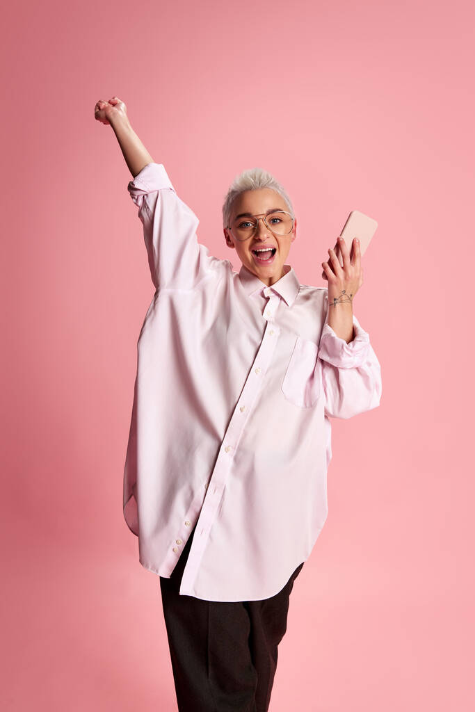Portret van jonge stijlvolle vrouw met kort haar poseren in wit shirt met telefoon geïsoleerd op roze achtergrond. Online winkelen. Concept van jeugd, schoonheid, mode, levensstijl, emoties, gezichtsuitdrukking - Foto, afbeelding