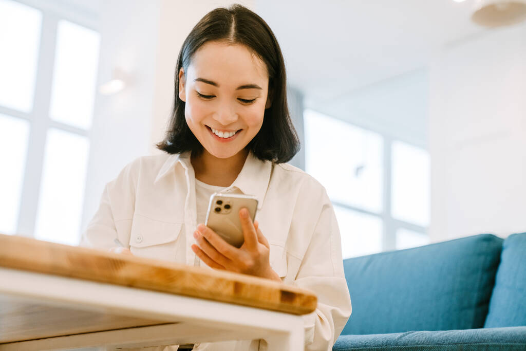 Joven linda sonriente mujer asiática sosteniendo el teléfono y mirando en la pantalla en acogedora sala de estar en casa - Foto, Imagen