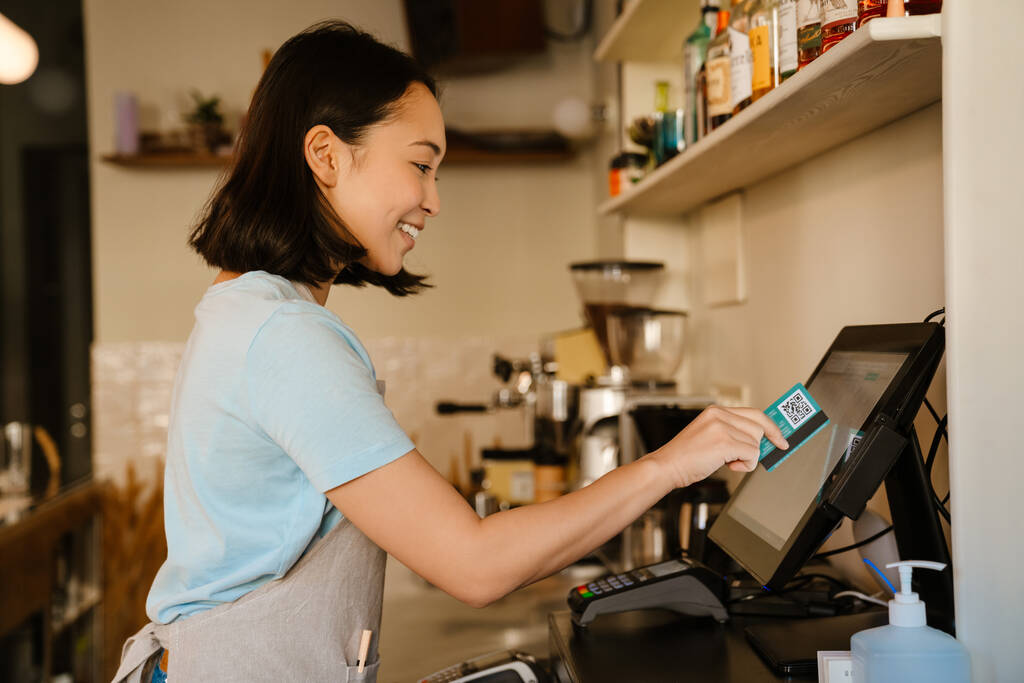 Ασιάτισσα μπαρίστα που φοράει ποδιά χαμογελώντας ενώ δουλεύει με ταμειακή μηχανή στο cafe - Φωτογραφία, εικόνα