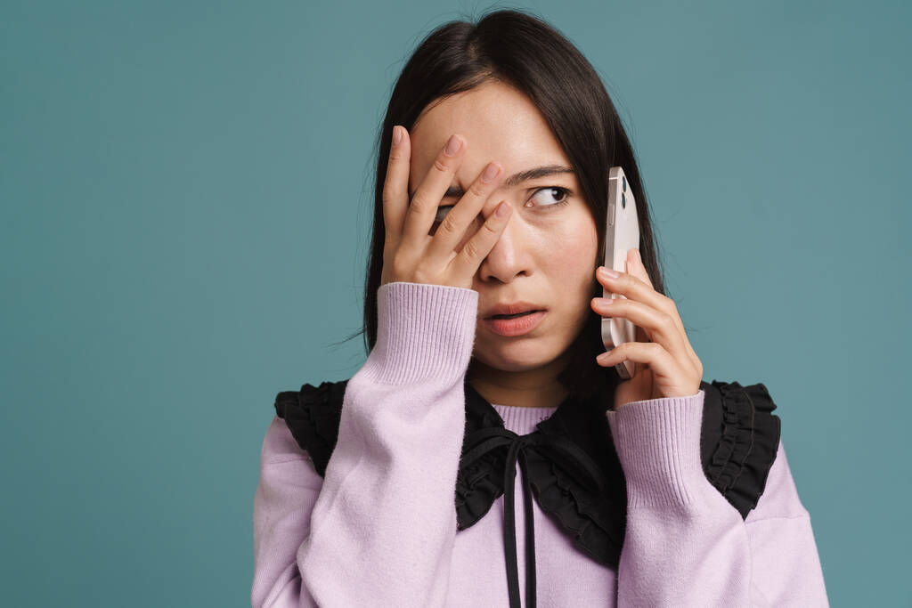 Jovem mulher asiática cobrindo seu rosto e falando no celular isolado sobre fundo azul - Foto, Imagem