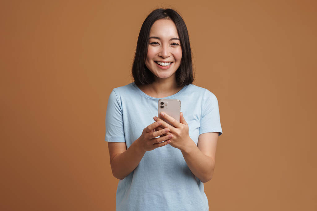 Νεαρή Ασιάτισσα γυναίκα που φοράει t-shirt γελώντας ενώ χρησιμοποιεί κινητό τηλέφωνο απομονωμένο σε μπεζ φόντο - Φωτογραφία, εικόνα