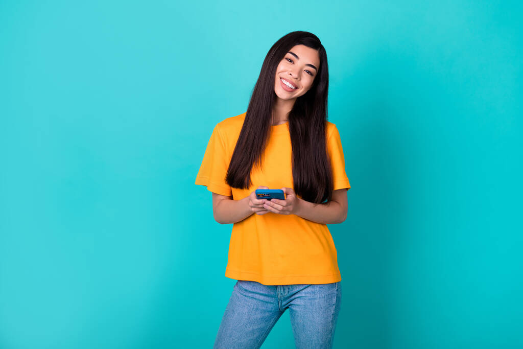 Fotografie optimistické mladé dámy držet telefon nosit oranžové tričko džíny izolované na pozadí teal barva. - Fotografie, Obrázek