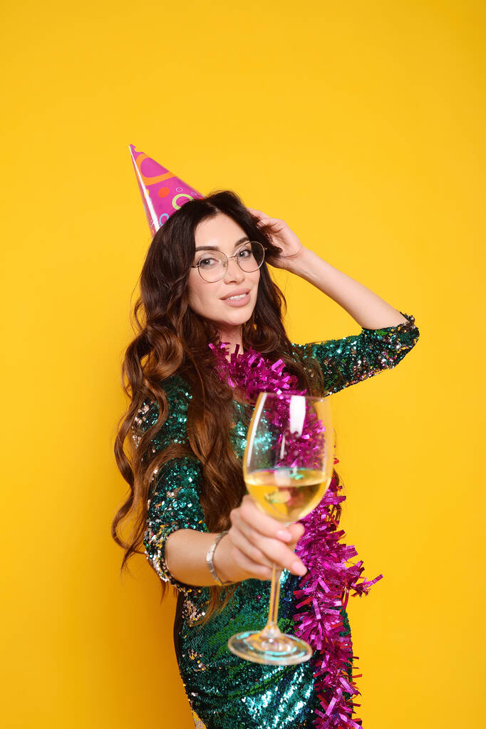 pretty young woman in a smart dress in a party cap with a pink boa around her neck and a glass of champagne smiling on a yellow background - Photo, Image