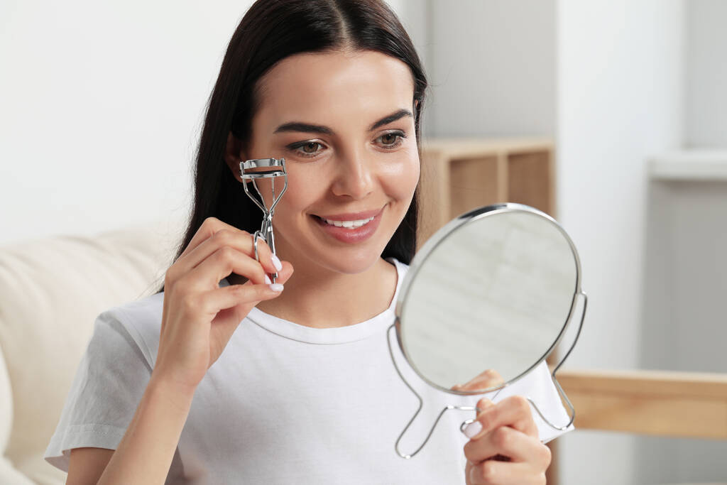 Όμορφη νεαρή γυναίκα με καθρέφτη χρησιμοποιώντας eyelash curler σε εσωτερικούς χώρους - Φωτογραφία, εικόνα