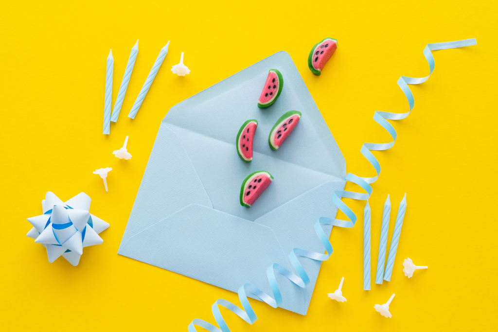 Top view of sweets on envelope near blue serpentine and festive candles on yellow background  - Photo, Image