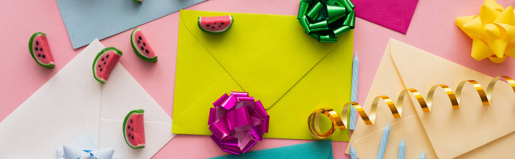 Vue du dessus des bonbons et des noeuds cadeaux sur des enveloppes colorées sur fond rose, bannière  - Photo, image