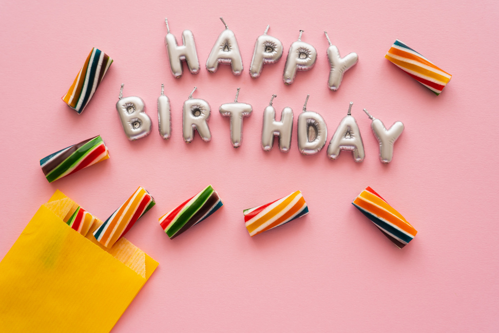 Vista superior de las velas en forma de letras de Feliz Cumpleaños cerca de dulces rayados sobre fondo rosa  - Foto, imagen
