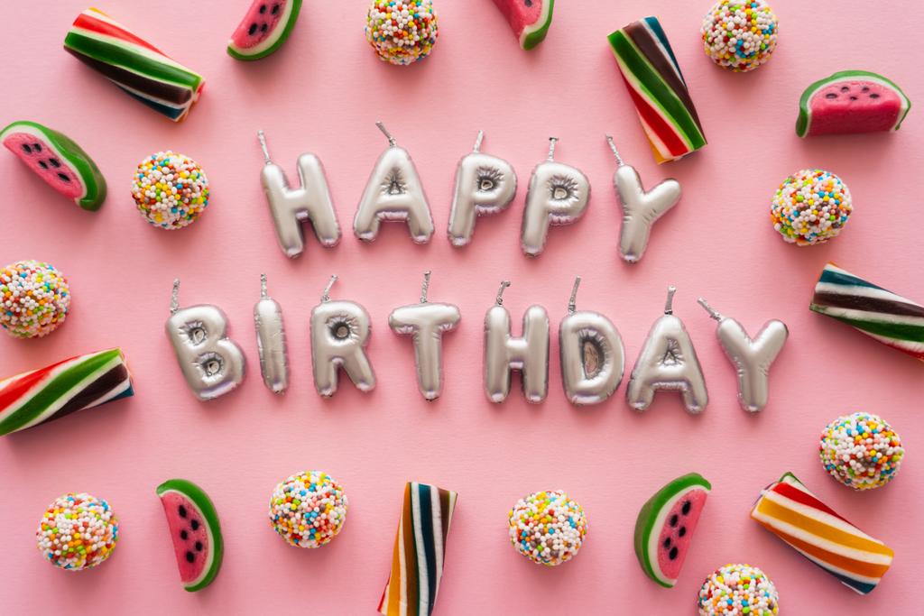 Vista dall'alto di caramelle colorate vicino a candele a forma di Happy Birthday lettering su sfondo rosa  - Foto, immagini