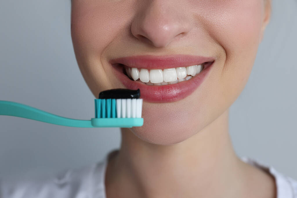 Vrouw met tandenborstel met houtskool tandpasta op grijze achtergrond, close-up - Foto, afbeelding