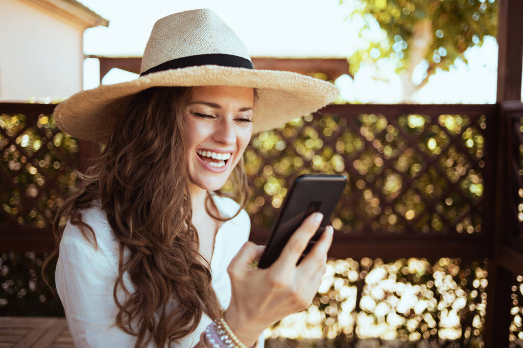 sorridente alla moda 40 anni casalinga in camicia bianca con cappello avendo video incontro su uno smartphone nella terrazza dell'hotel guest house. - Foto, immagini