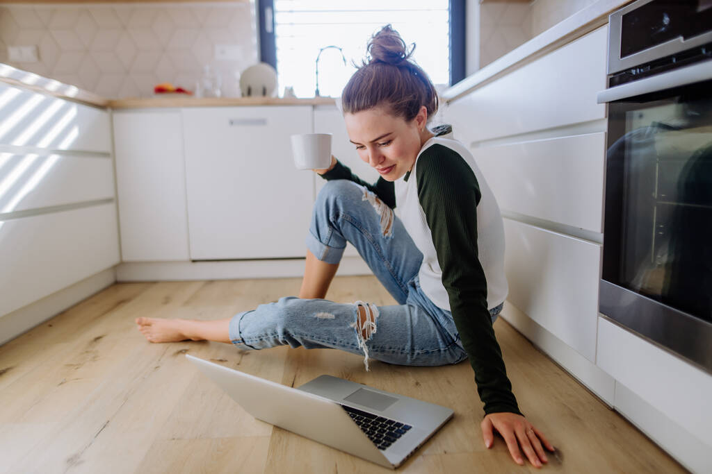 Młoda kobieta siedzi z laptopem i filiżanką kawy w kuchni. - Zdjęcie, obraz