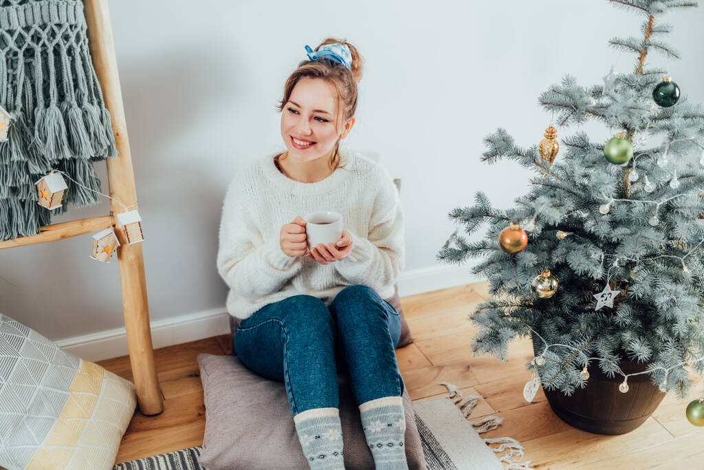 居心地の良いセーターを着た幸せな若い女性は、モダンな北欧のインテリアの家で鉢植えのクリスマスツリーの近くの床のクッションでリラックスし、熱いお茶を飲んでいます。環境に優しい居心地の良い冬の休日。選択的焦点 - 写真・画像