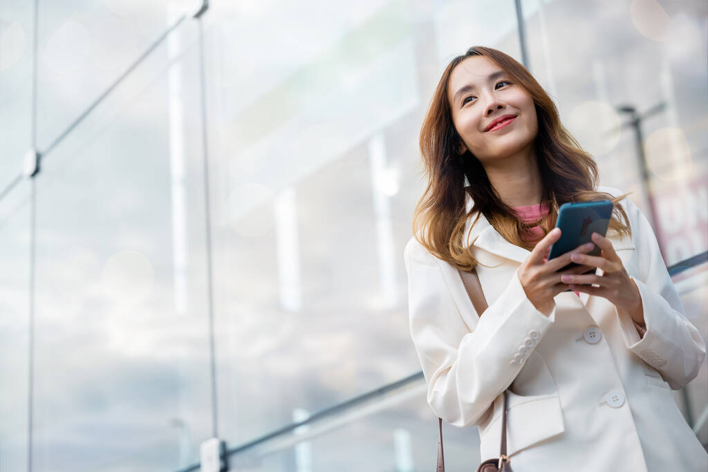 Πορτρέτο επιτυχημένη επιχειρηματίας χαμογελώντας κρατώντας smartphone χρήση chat εφαρμογή σε απευθείας σύνδεση το πρωί, Ασίας επιχειρηματίας που εργάζονται με κινητό τηλέφωνο στέκεται κατά δρόμο κτίριο μπροστά κοντά στο γραφείο - Φωτογραφία, εικόνα