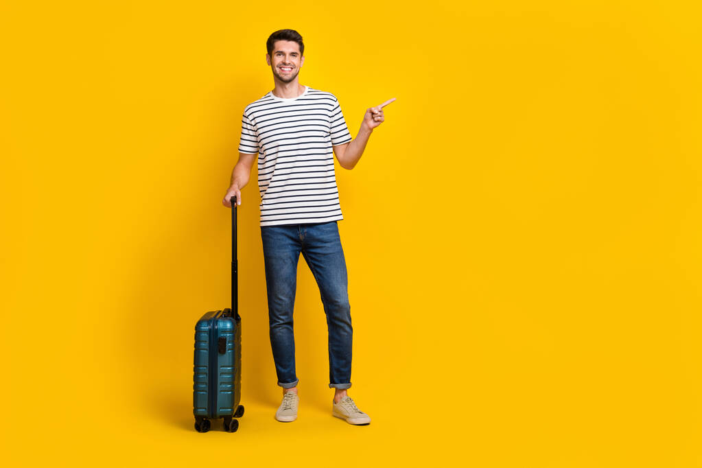 Full length φωτογραφία του καλού άντρα διάθεση ντυμένος ριγέ t-shirt δείχνοντας κενό χώρο κρατώντας αποσκευές απομονωμένο κίτρινο χρώμα φόντο. - Φωτογραφία, εικόνα