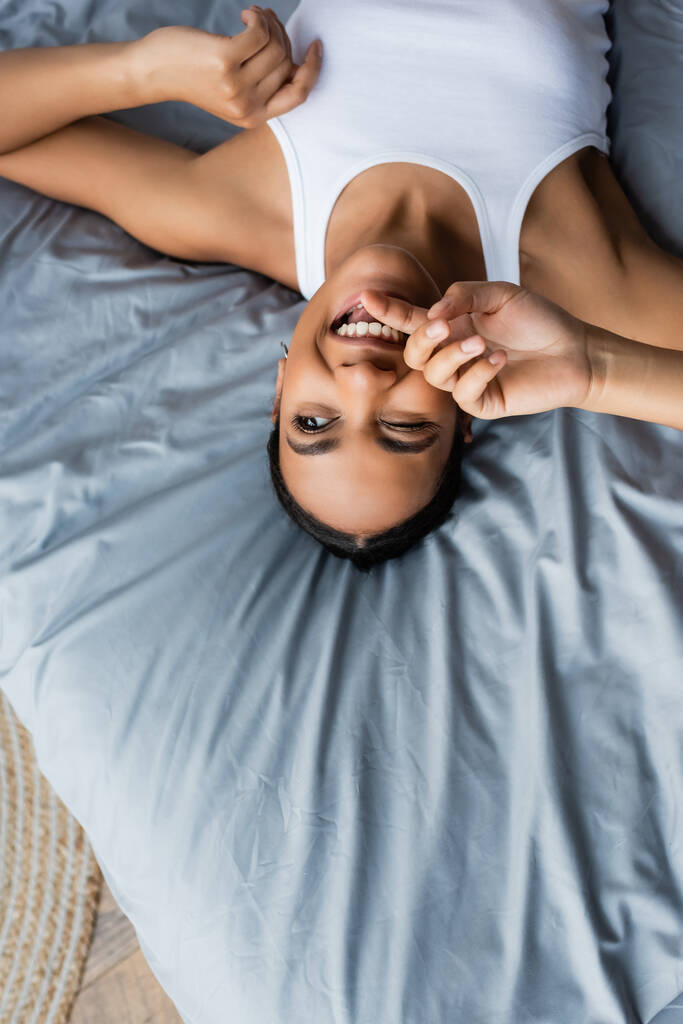 vista superior de la alegre mujer afroamericana guiñando un ojo mientras está acostada en la cama en casa - Foto, Imagen