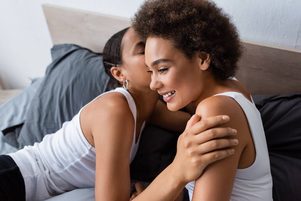 Αφροαμερικανή λεσβία που φιλάει μάγουλο ευτυχισμένης κοπέλας στην κρεβατοκάμαρα.  - Φωτογραφία, εικόνα