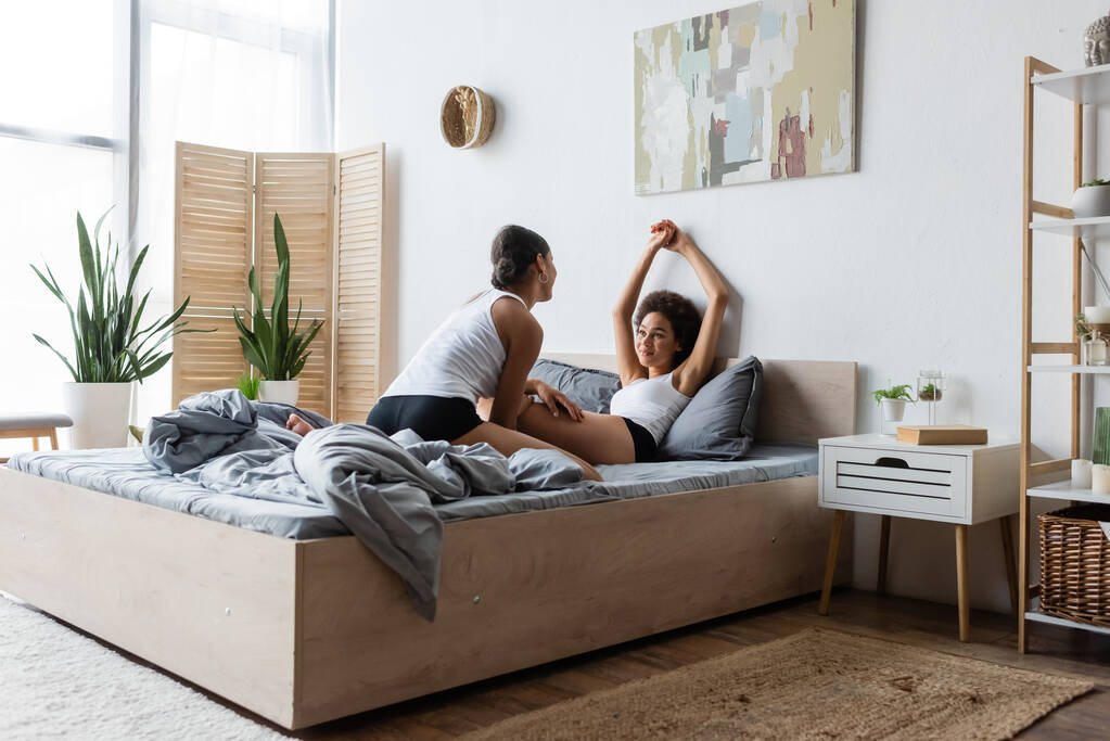 junge lesbische Afroamerikanerin schaut glückliche Freundin mit erhobenen Händen im modernen Schlafzimmer an - Foto, Bild