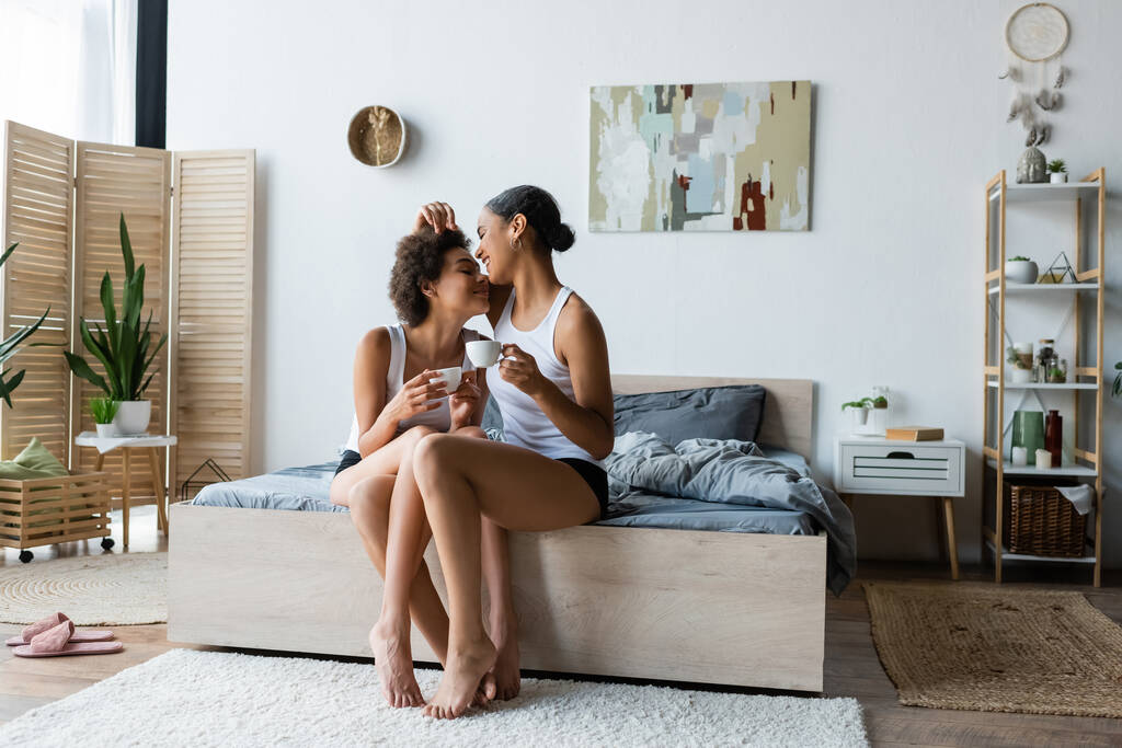full length of cheerful lesbian african american couple holding cups of coffee while sitting on bed - Photo, Image