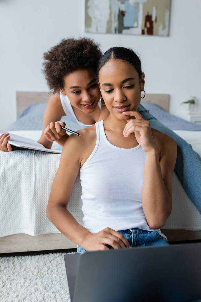 Kıvırcık Afrikalı Amerikalı kadın dizüstü bilgisayarıyla yatak odasındaki kız arkadaşına işaret ediyor. - Fotoğraf, Görsel