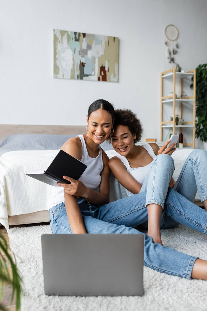 glückliche und lesbische afrikanisch-amerikanische Frauen schauen auf Laptop im Schlafzimmer  - Foto, Bild