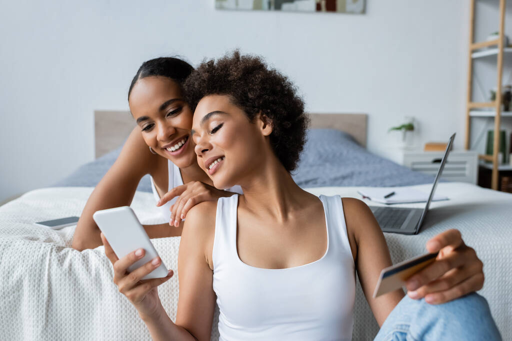 幸せなアフリカ系アメリカ人レズビアン女性スマートフォンを使ってクレジットカードを持ちながら彼女の近くに - 写真・画像
