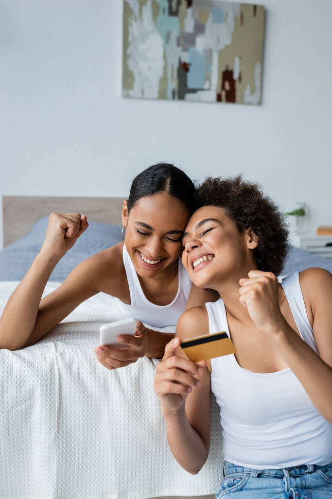 glücklich afrikanisch-amerikanische lesbische Frau hält Kreditkarte nahe aufgeregter Freundin mit Smartphone  - Foto, Bild