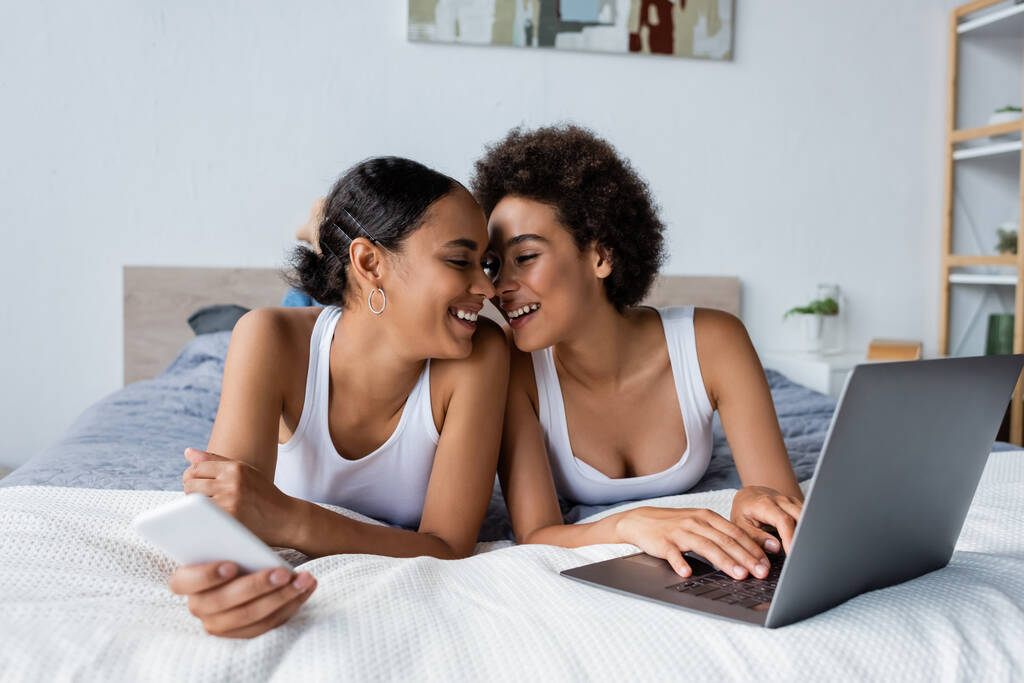 feliz africano americano lesbiana pareja sonriendo cerca laptop en cama - Foto, Imagen