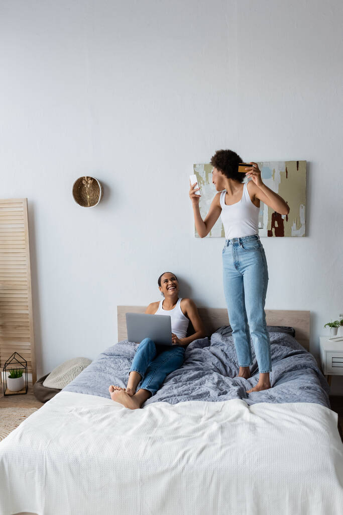 izgatott afro-amerikai leszbikus nő gazdaság smartphone és hitelkártya, miközben áll az ágyban közelében barátnő   - Fotó, kép