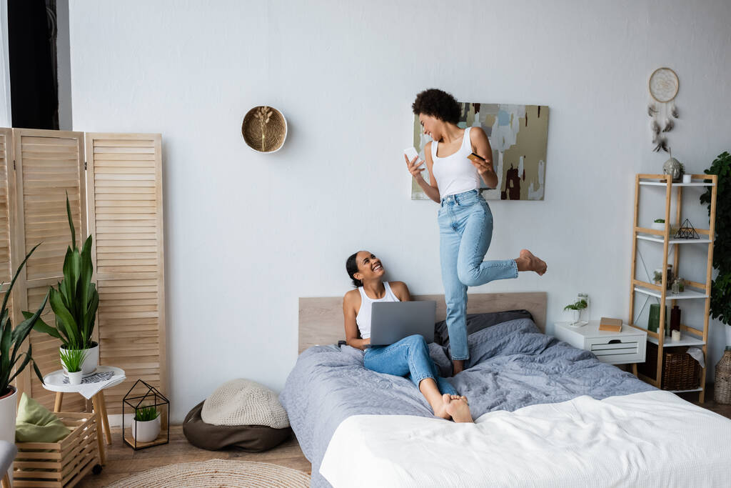 eccitata donna lesbica africana americana in possesso di smartphone e carta di credito mentre in piedi sul letto vicino alla fidanzata sorridente   - Foto, immagini