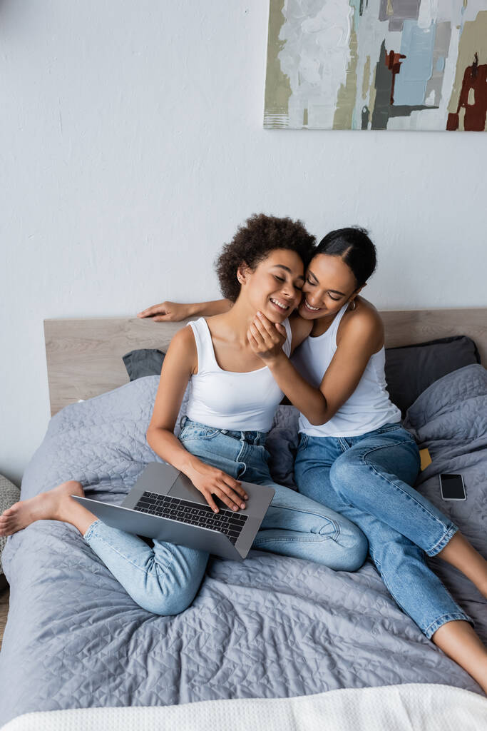 Afrikanisch-amerikanisches lesbisches Paar umarmt sich in der Nähe von Laptop und Smartphone auf dem Bett  - Foto, Bild