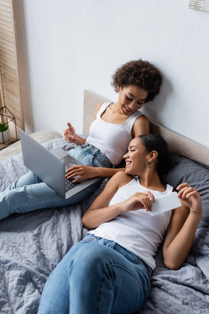 feliz mulher lésbica afro-americana com laptop conversando com namorada segurando smartphone no quarto  - Foto, Imagem