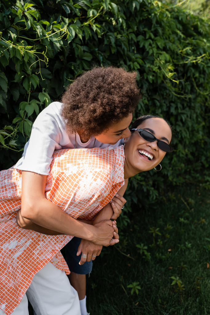 happy african american lesbian woman embracing cheerful girlfriend in stylish sunglasses laughing in park - Photo, Image