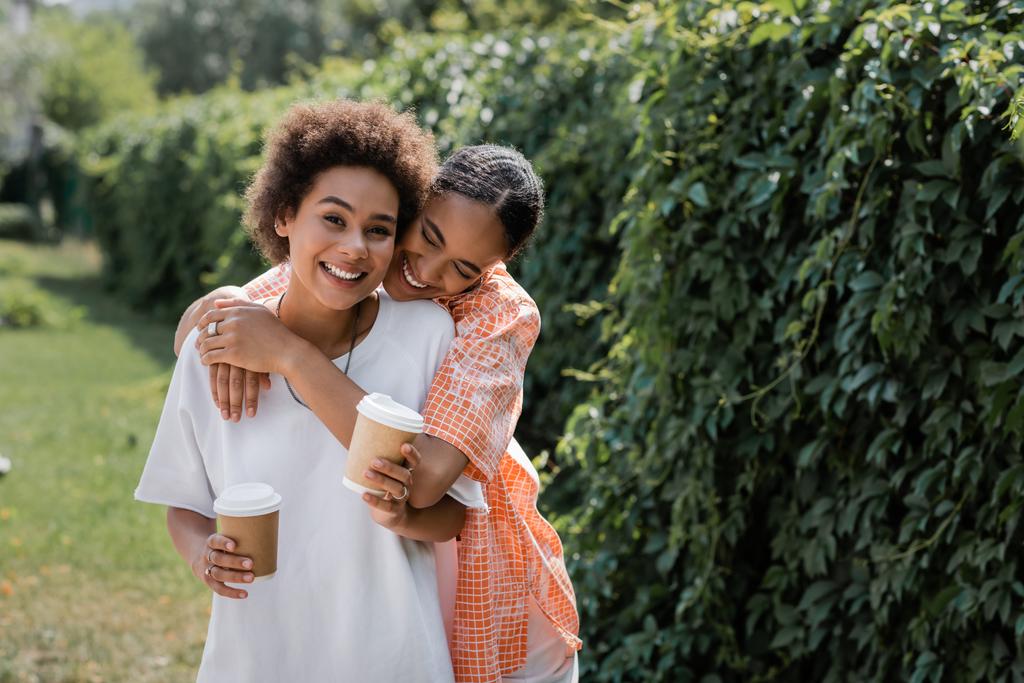 feliz casal lésbico afro-americano segurando copos de papel com café para ir enquanto abraça no parque  - Foto, Imagem