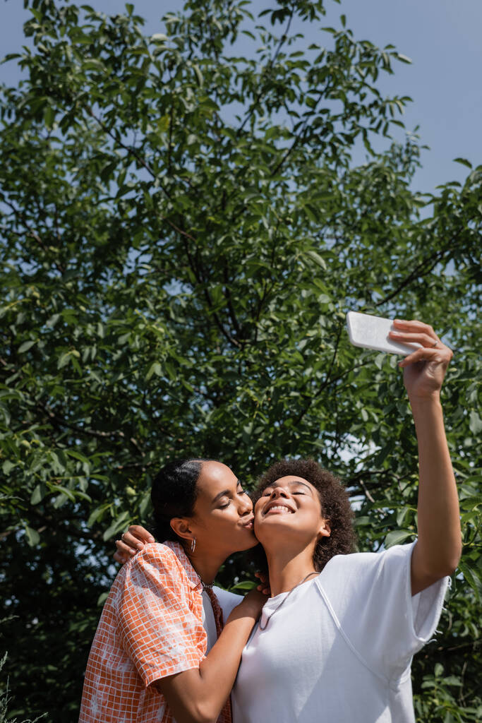 gelukkig afrikaans amerikaans lesbisch vrouw zoenen wang van vriendin terwijl het nemen van selfie in de buurt boom in park  - Foto, afbeelding
