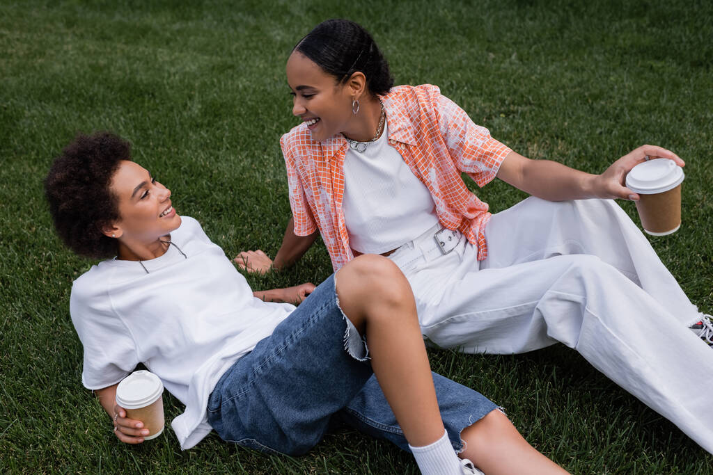 joyeuses femmes lesbiennes afro-américaines tenant des tasses en papier et reposant sur l'herbe dans le parc - Photo, image