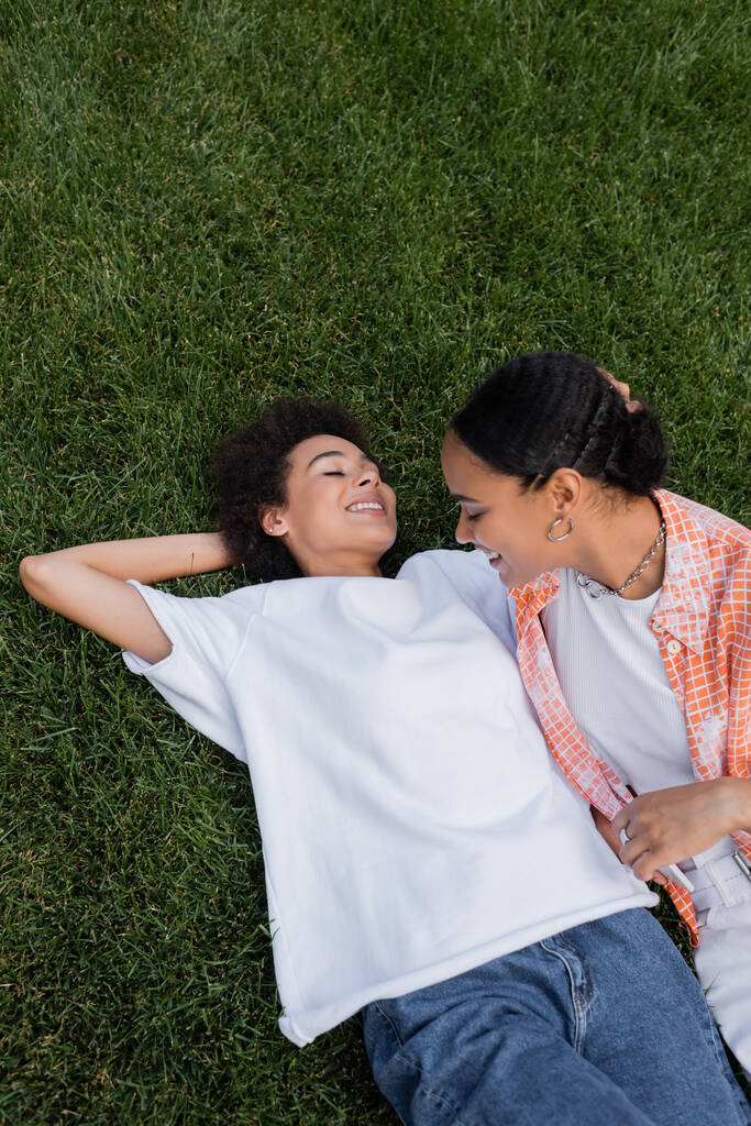 幸せなアフリカ系アメリカ人レズビアン女性のトップビュー彼女の近くにスマートフォンを持っている草の上に横たわる  - 写真・画像